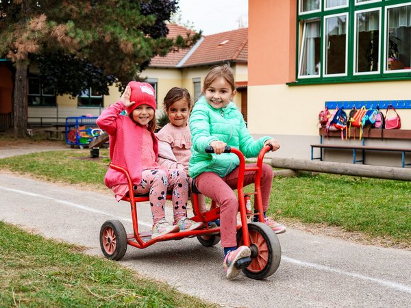 Drei Kinder fahren auf einem Dreirad, im Hintergrund ein Kindergartengebäude