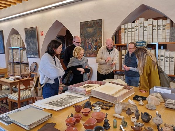 Gruppe von Menschen in einer historischen Bibliothek, die alte Dokumente und archäologische Funde betrachtet.