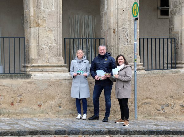 Drei Personen mit Informationsmaterial vor einer alten Steinmauer neben einer Bushaltestelle.