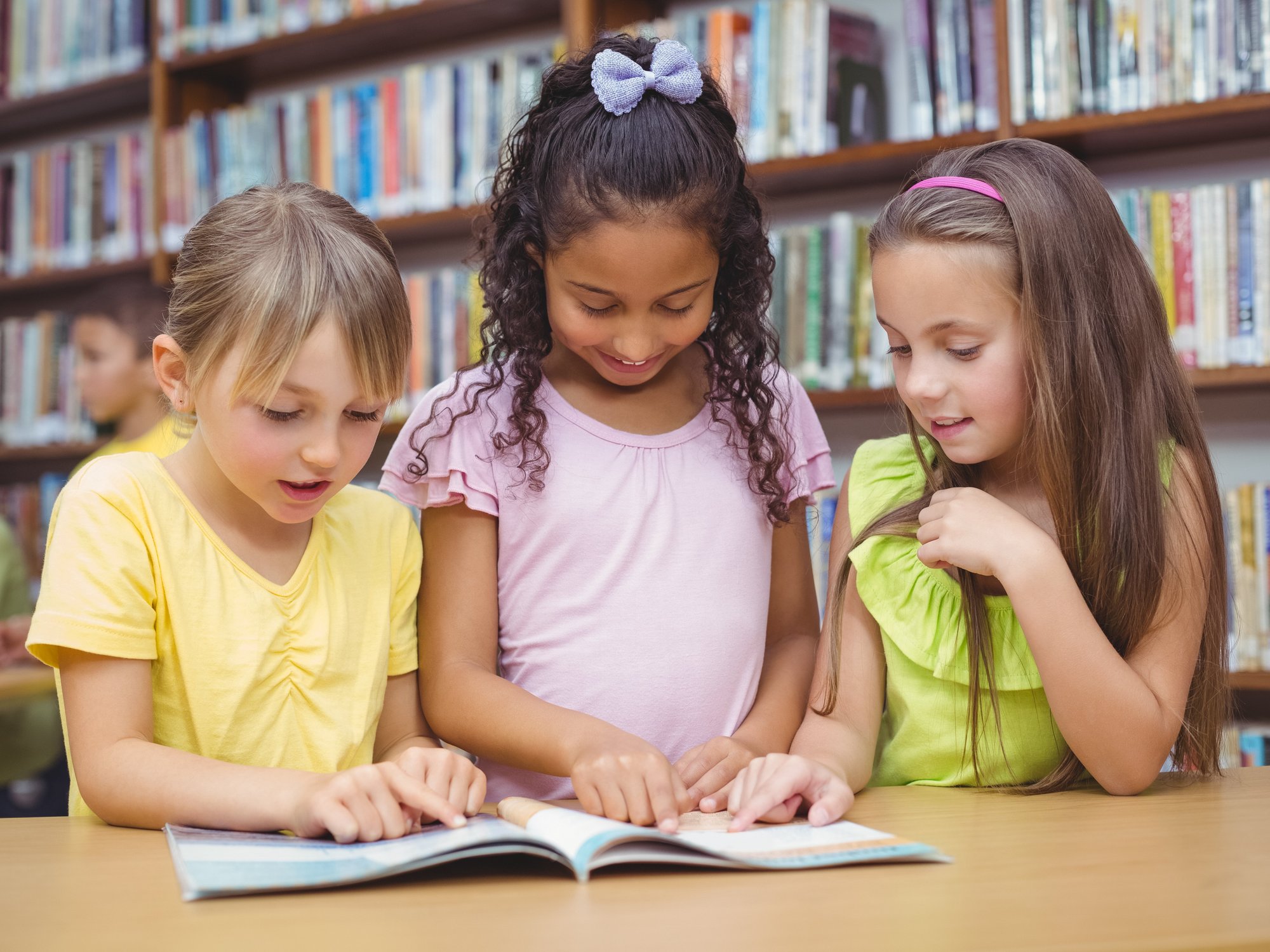 Drei Mädchen stehen nebeneinander an einem Tisch in  einer Bücherei und betrachten ein aufgeschlagenes Buch