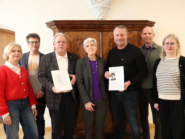 Gruppenfoto von sieben Personen vor einem Holzschrank, zwei Personen halten Dokumente mit Text und Porträtfotos.